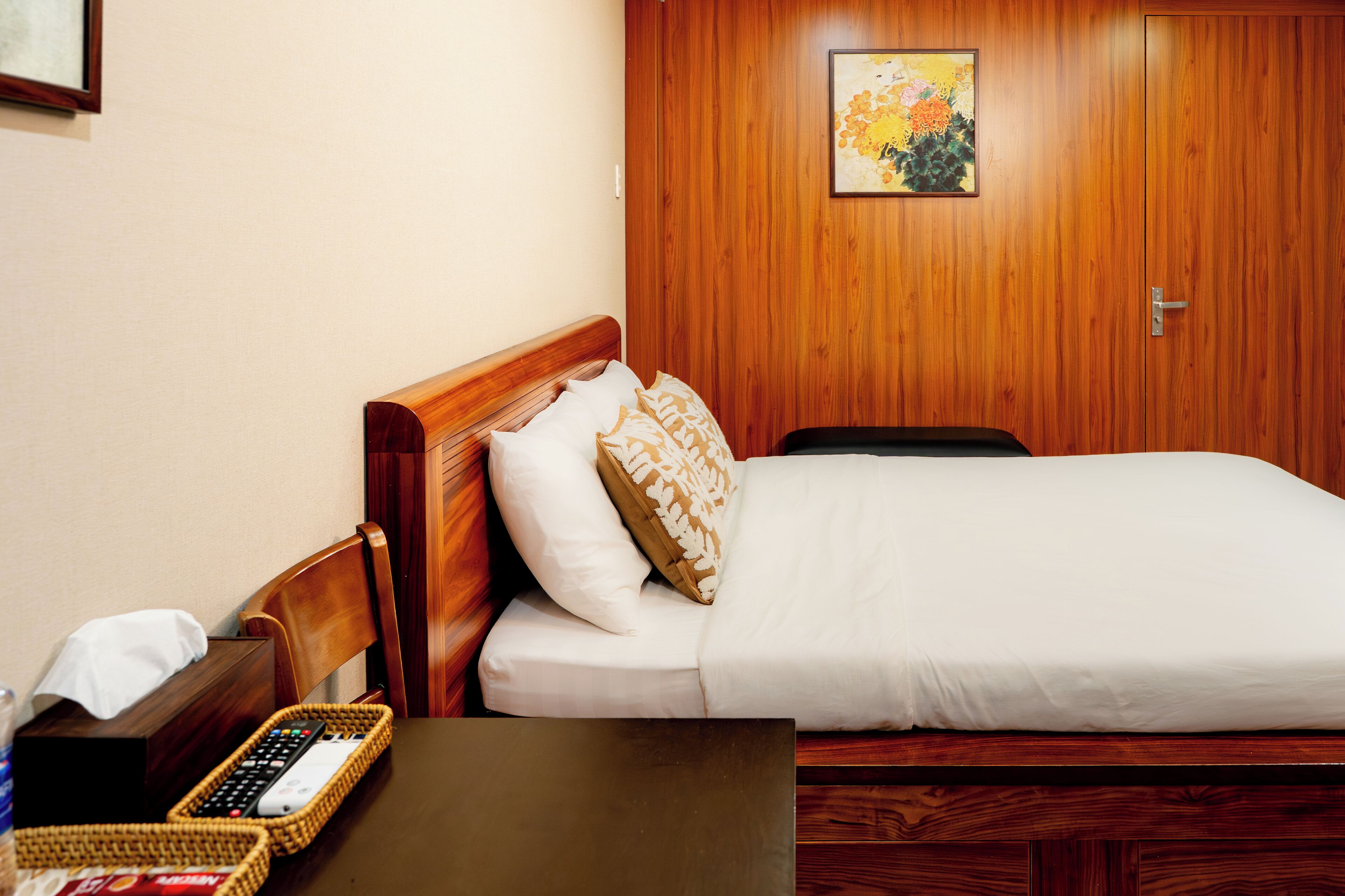 A hotel room with a wooden headboard, a bed with white linens, a wooden nightstand with a remote control, and a framed picture on the wall.
