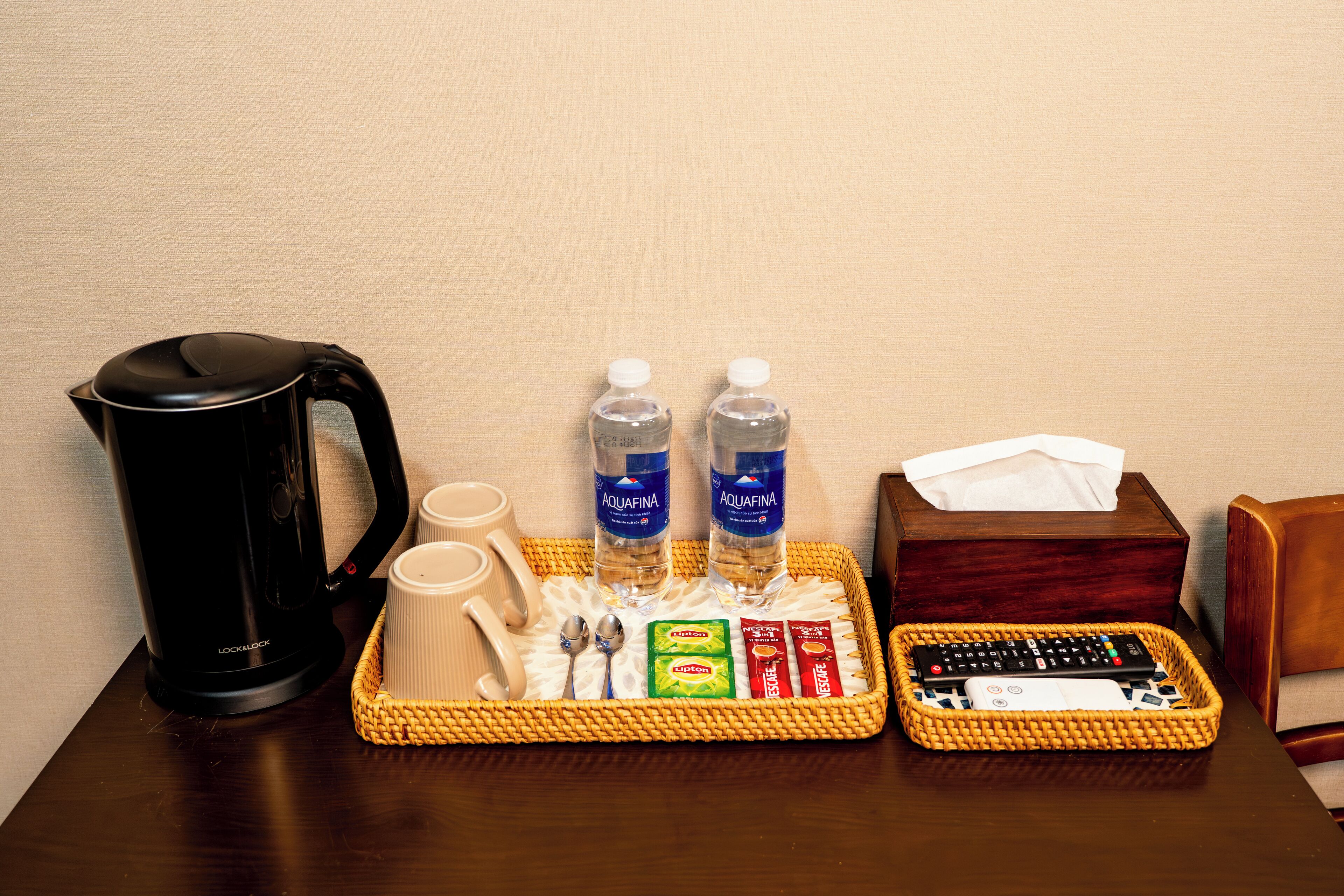 A tray with a coffee maker, two bottles of water, cups, spoons, condiments, and a remote control on a wooden table.