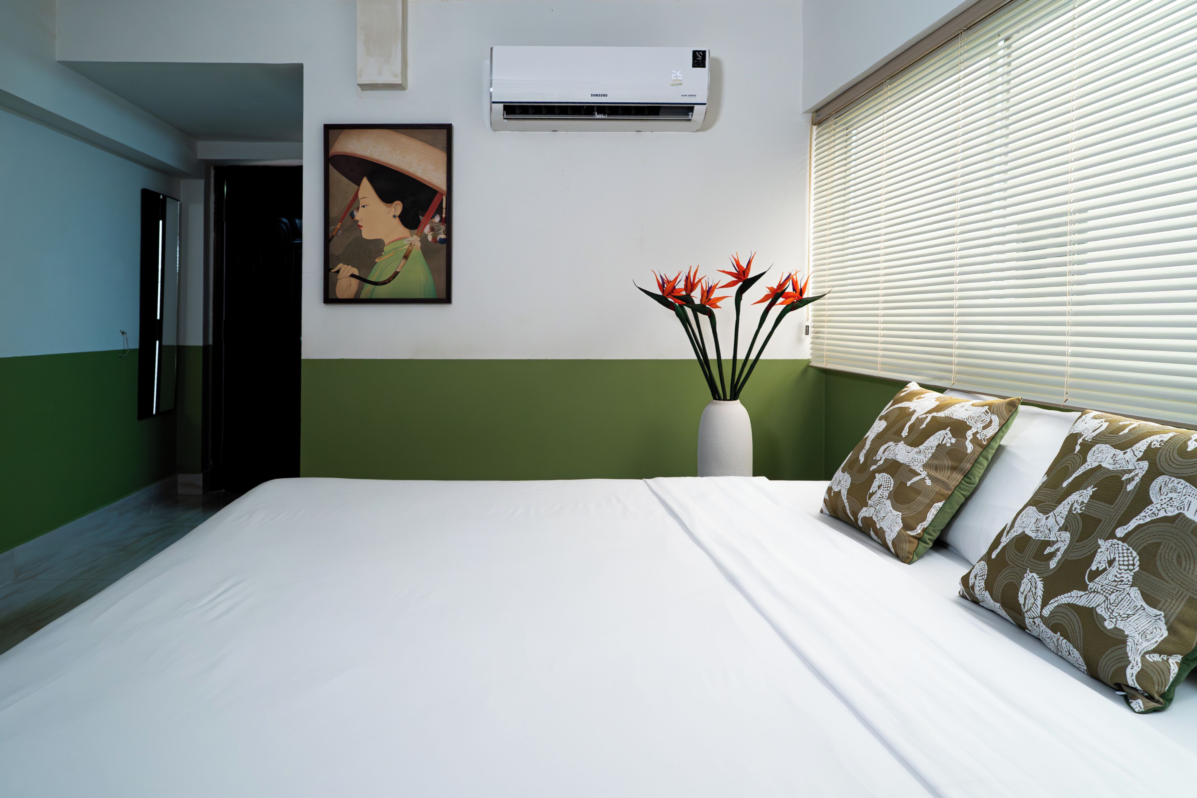 A hotel room with a bed, a painting on the wall, an air conditioner, a window with blinds, and a vase with flowers.