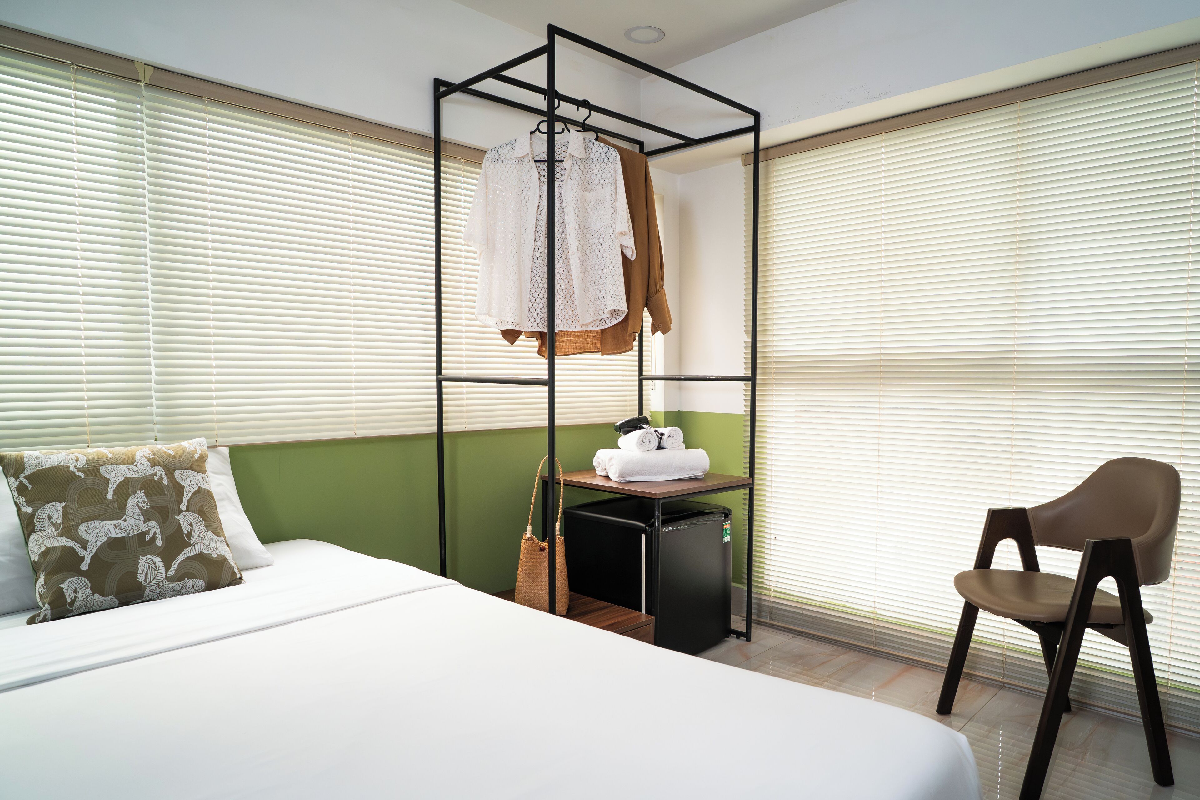A hotel room with a bed, a small table with a chair, a hanging wardrobe, and a window with blinds.