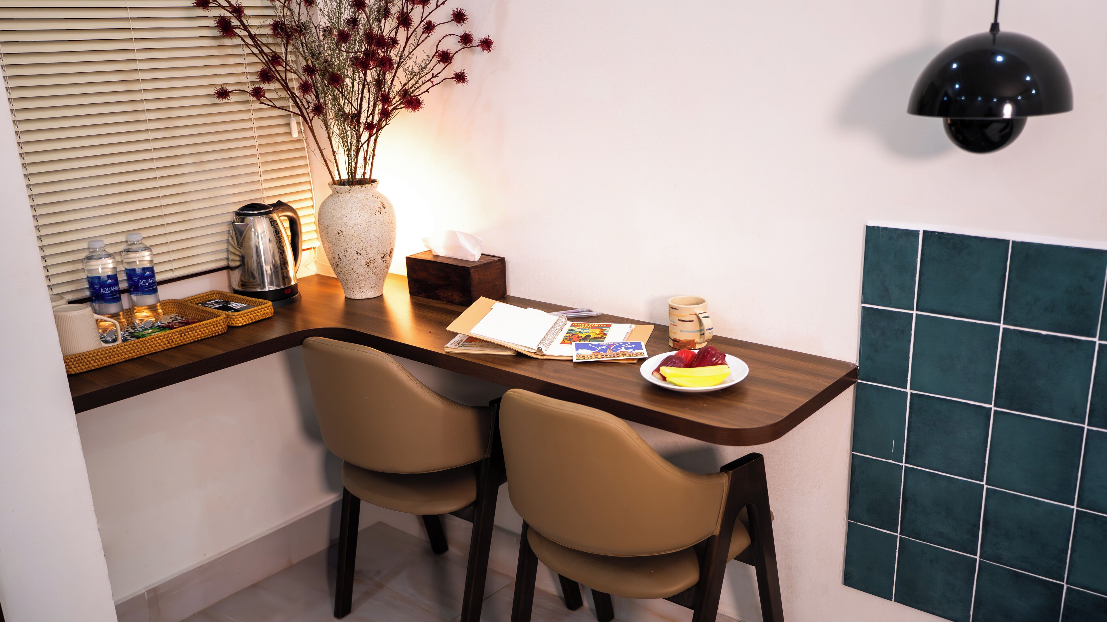 A small dining area with a wooden table, two chairs, a vase with dried flowers, a kettle, water bottles, a coffee maker, a notebook, a mug, and a plate with fruit.