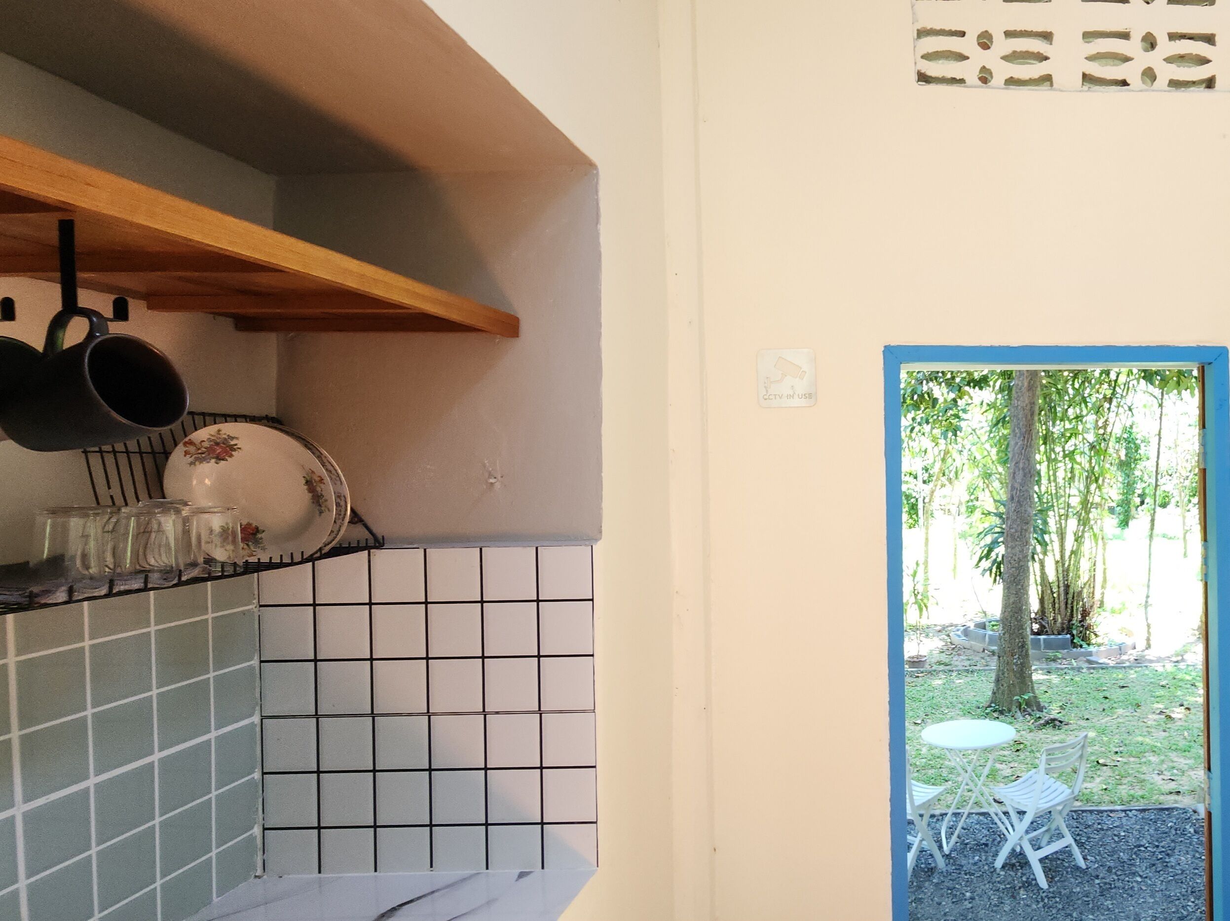 A kitchen with a wooden shelf, a tiled backsplash, and a door leading to an outdoor seating area with a tree and a table.