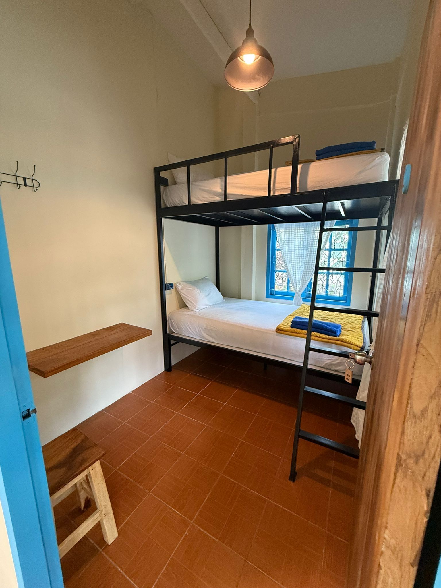 A bunk bed room with a hanging light fixture, a small wooden stool, and a window with blue shutters.