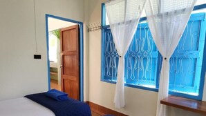 A room with two beds, a wooden door, and a window with blue metal bars and white curtains. - Baan Hinlad Home and Hostel (Koh Samui)
