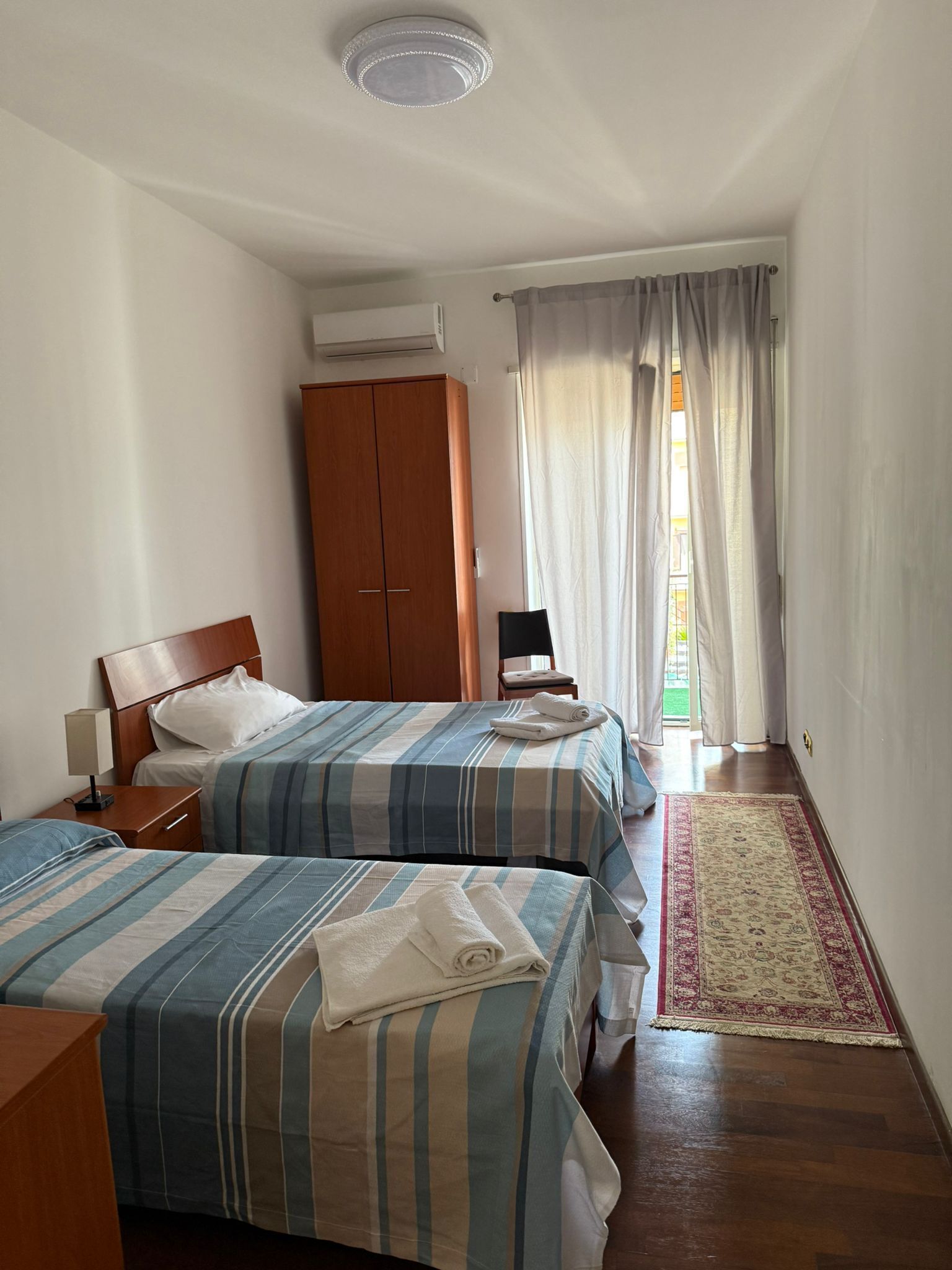 A hotel room with two beds, a wardrobe, a desk, and a window with curtains.
