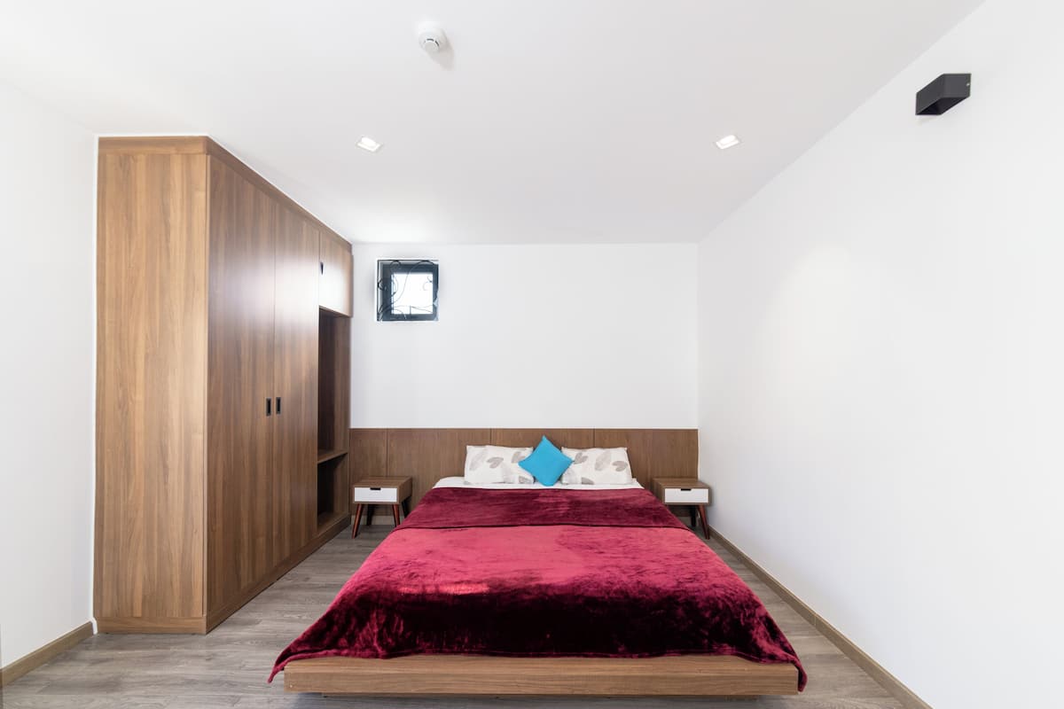 A bedroom with a wooden wardrobe, a bed with a red blanket, two bedside tables, and a small window.