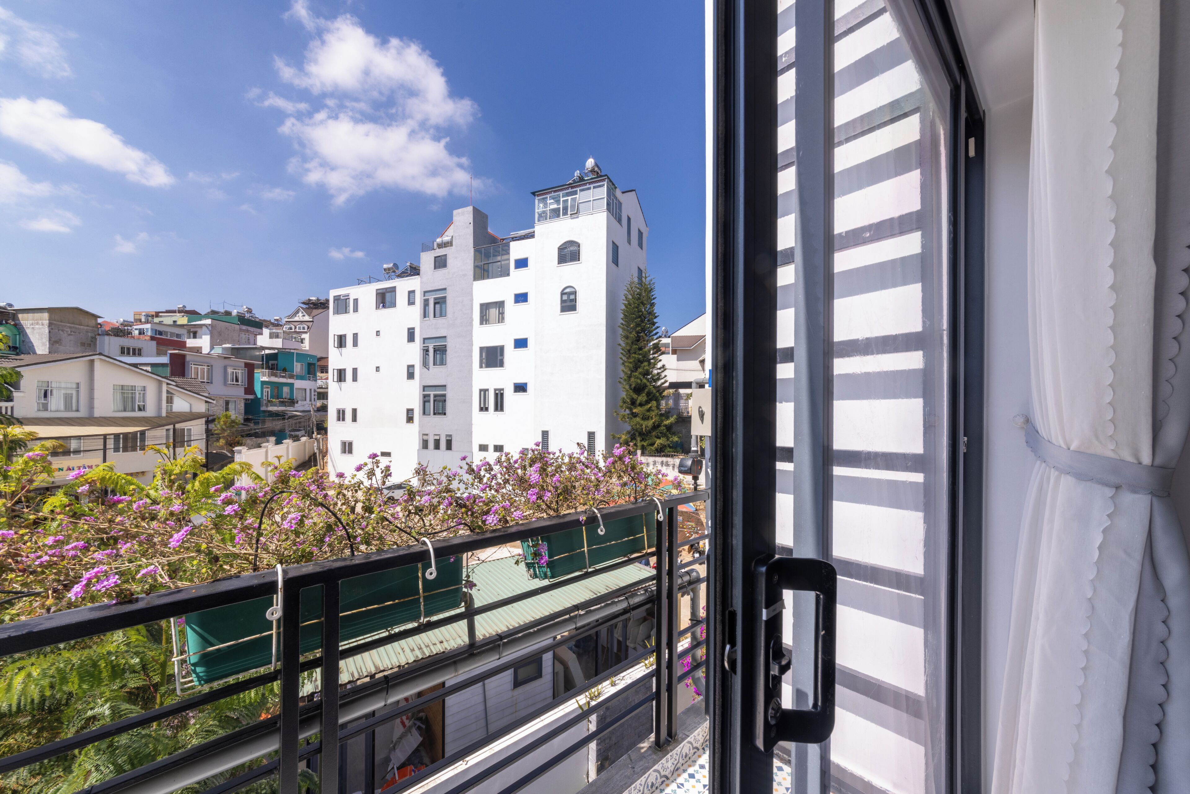 A view from a window overlooking a building with multiple floors, a balcony with a green roof, and a clear blue sky.