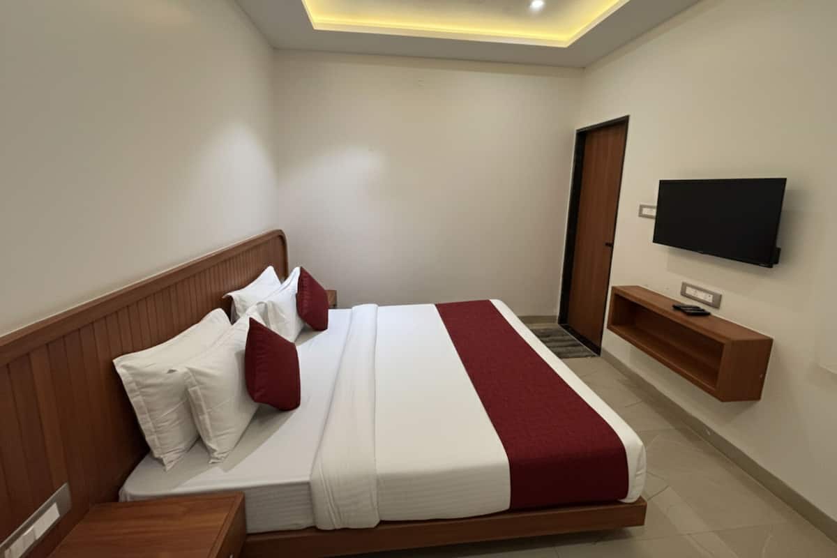 A modern hotel room with a large bed, wooden headboard, white and red bedding, a flat-screen TV, and a small wooden side table.