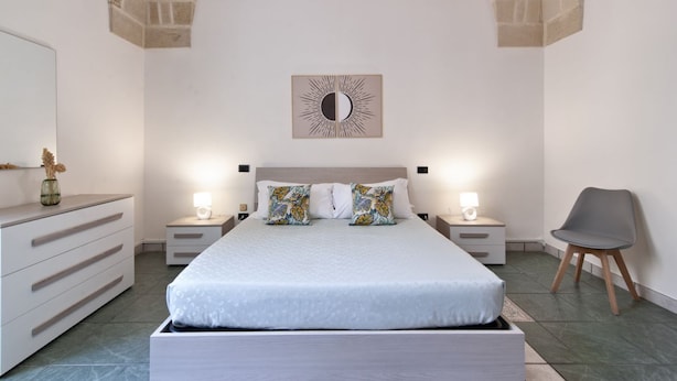 A bedroom with a large bed, two bedside tables with lamps, a dresser, a chair, and a wall-mounted mirror. - Sardinia 10 - Lecce Selection (Lecce)