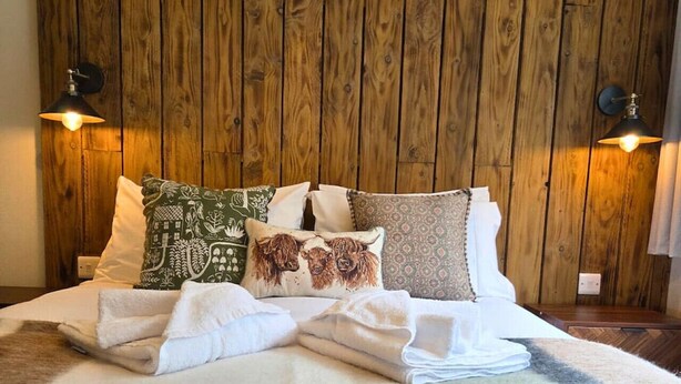 Una cama bien hecha con almohadones decorativos y un edredón esponjoso, frente a una pared de madera con dos lámparas de pared.