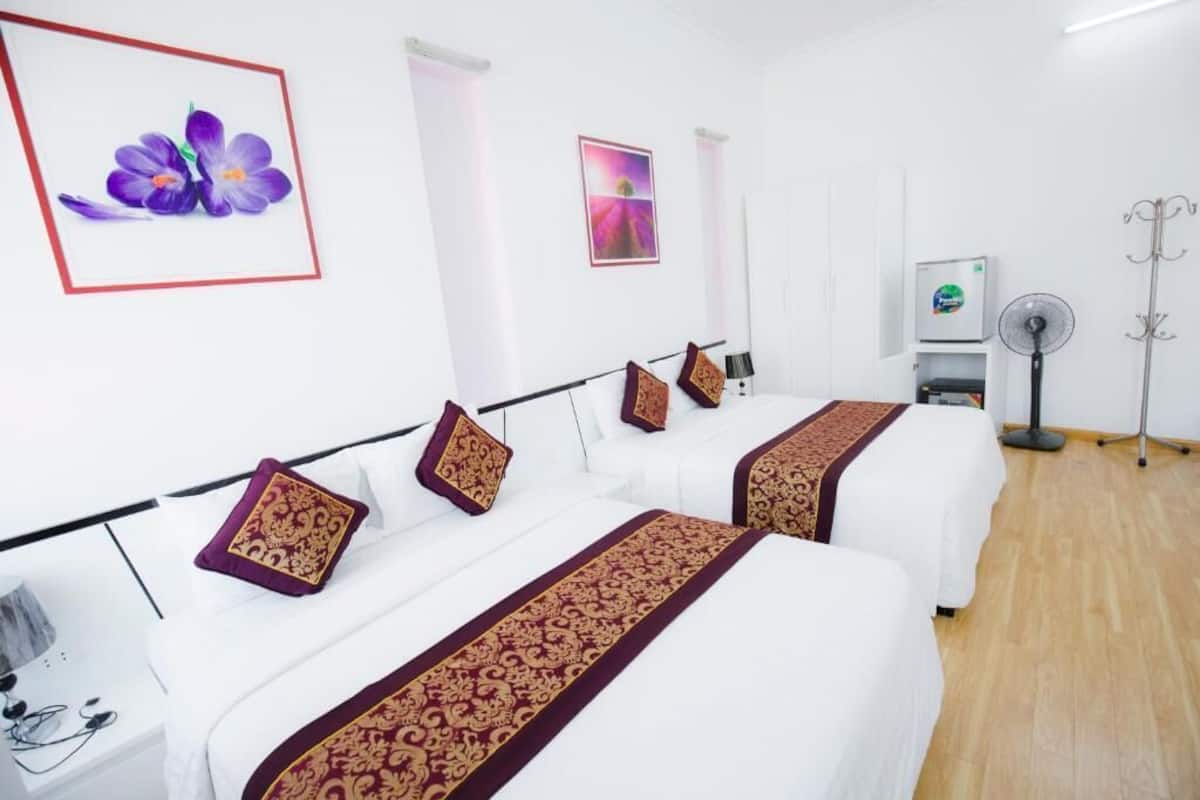 A hotel room with two beds, each with a white bedspread and a red and gold patterned runner. There are decorative pillows, a framed picture of flowers on the wall, and a standing fan in the corner.