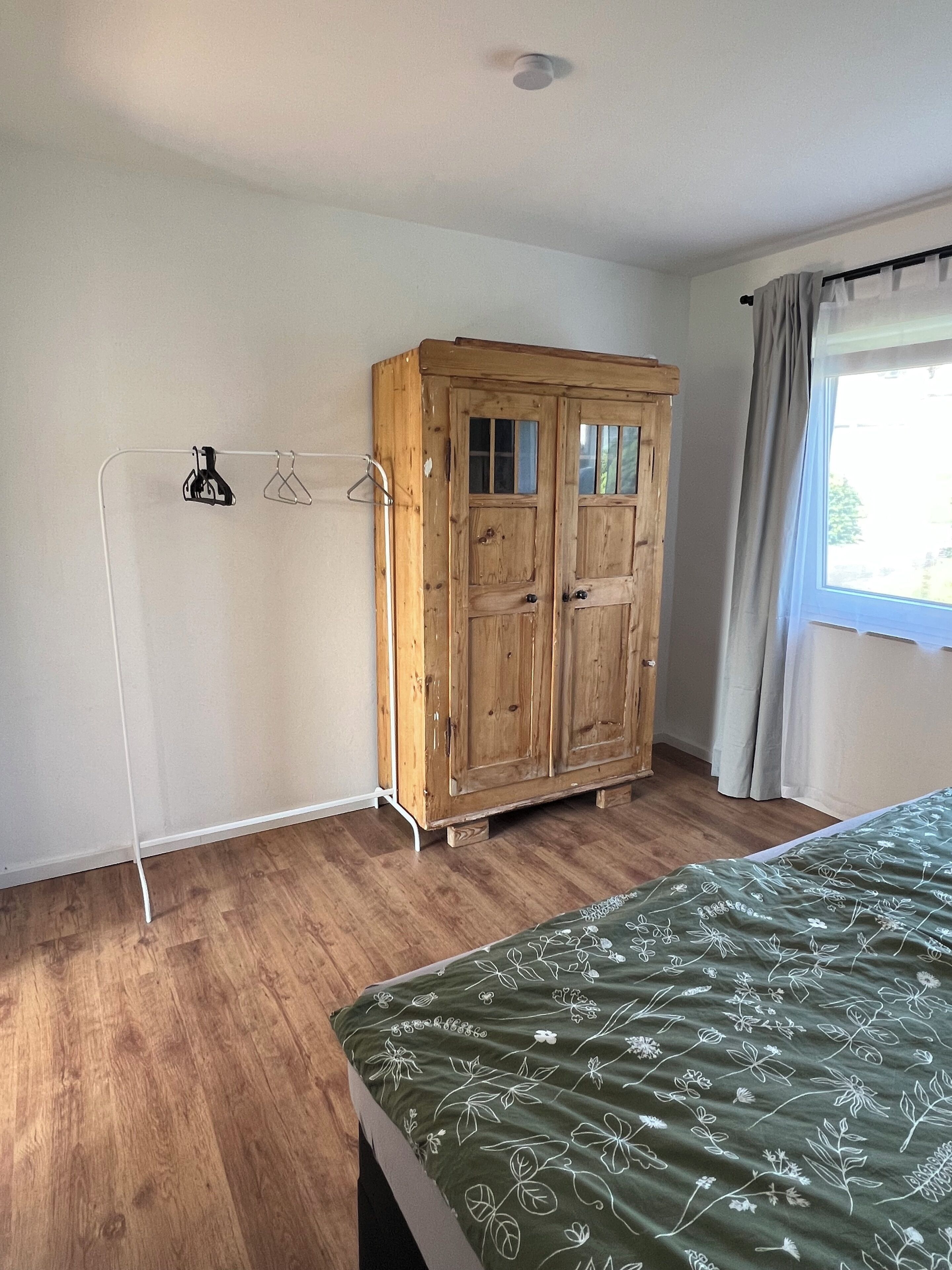 A bedroom with a wooden wardrobe, a bed with a floral bedspread, and a window with curtains.