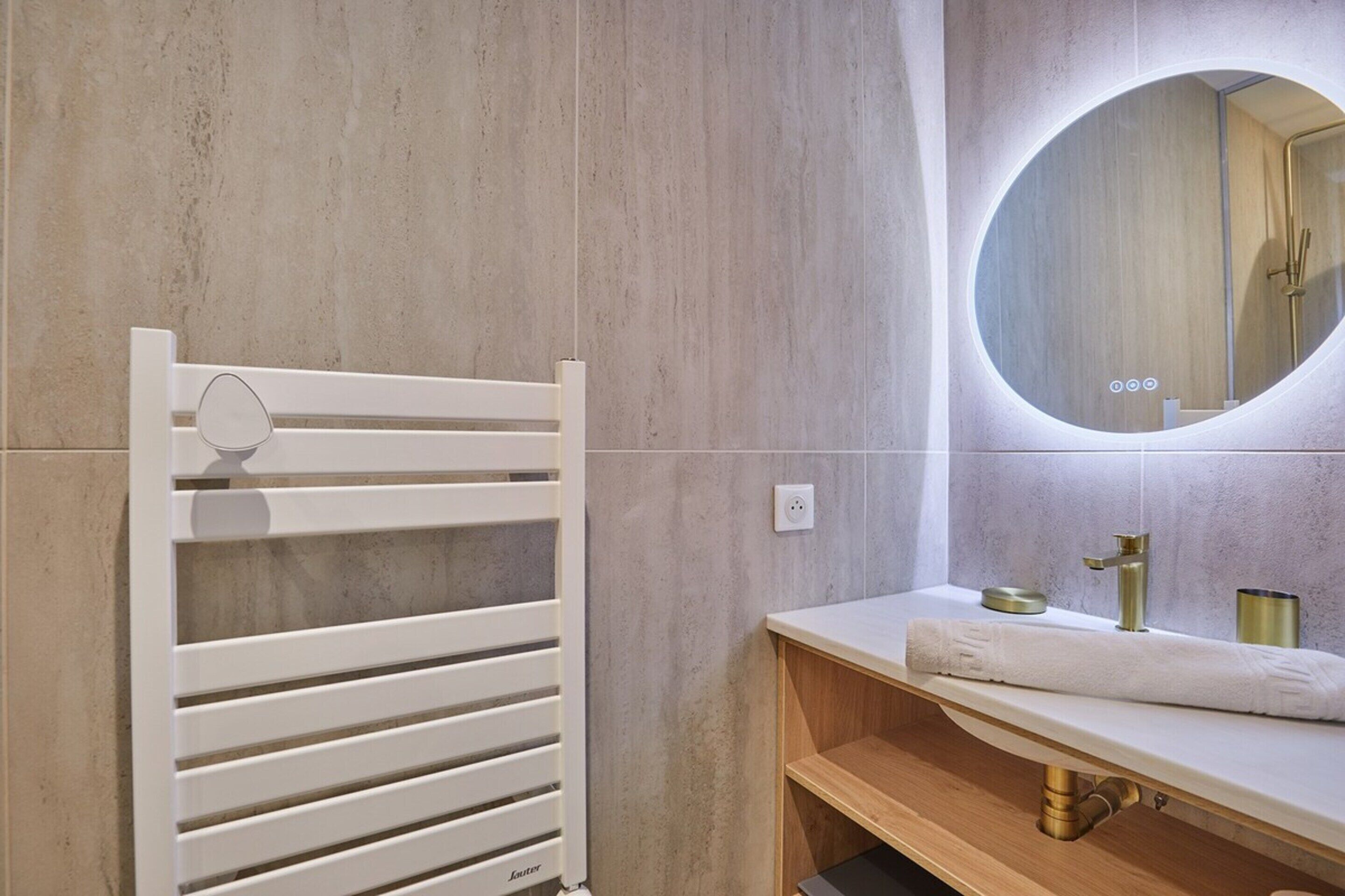 A modern bathroom with a white towel rack, a round mirror, and a wooden vanity with a gold faucet.