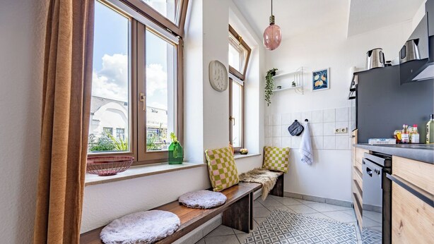 Une cuisine avec un banc intégré, une fenêtre avec des rideaux, un luminaire suspendu et un sol carrelé.