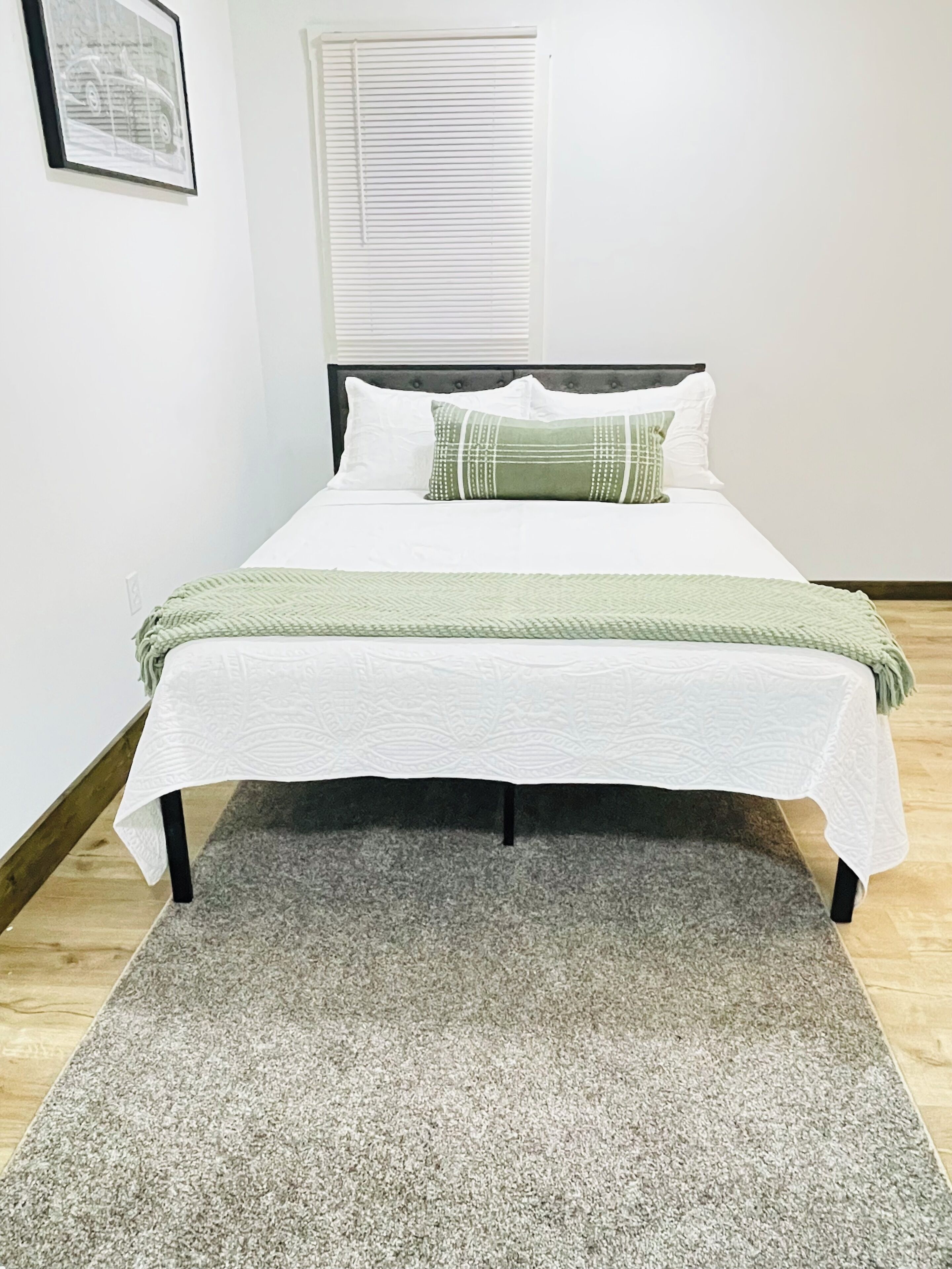 A neatly made bed with a white bedspread and a green pillow, a grey rug, and a window with blinds in the background.