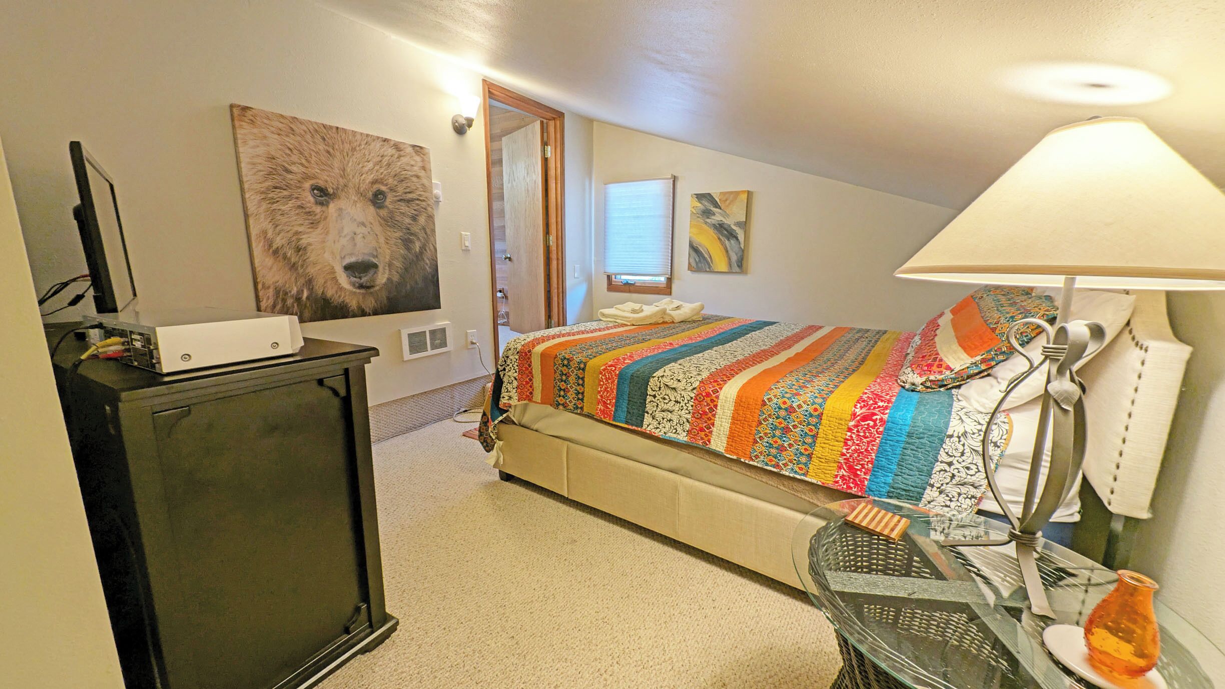 A bedroom with a bed, a desk, a lamp, a painting, and a window.