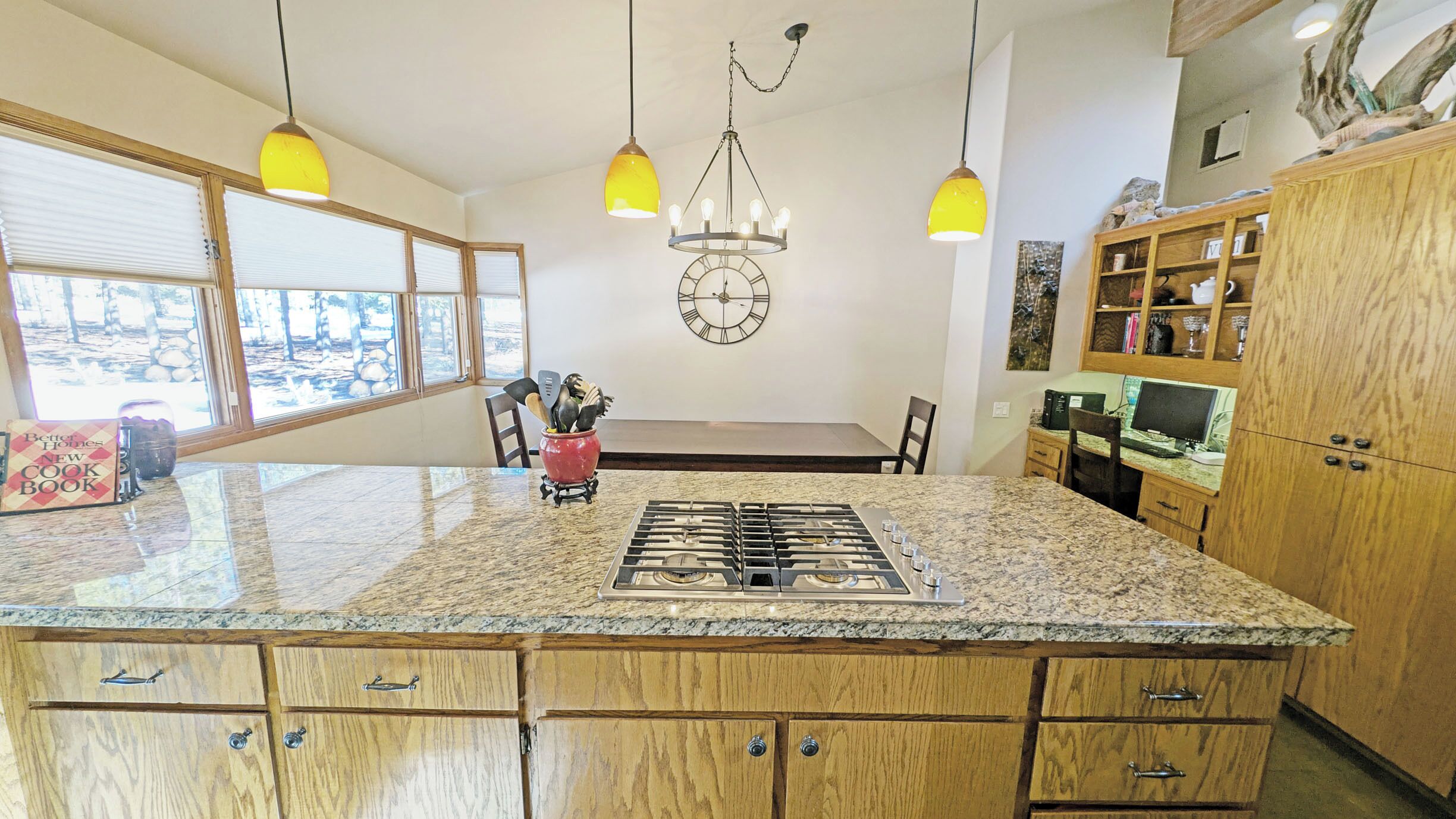 Una cocina con encimeras de granito, armarios de madera, una isla central con estufa a gas, luces colgantes y un reloj de pared.