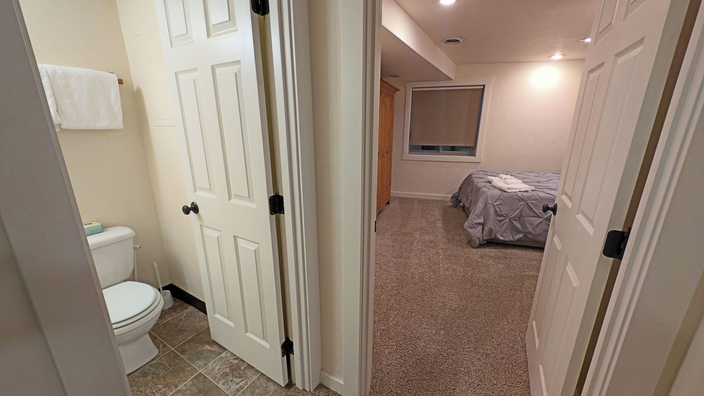 A bathroom with a toilet, a towel hanging on the wall, and a door leading to a bedroom with a bed and a window.