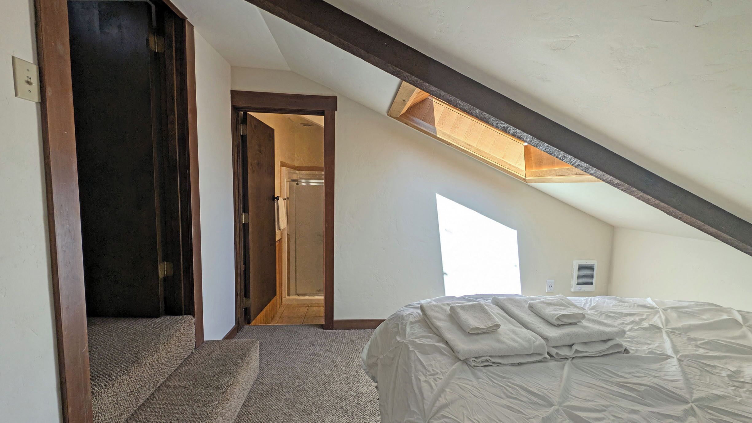Una camera da letto con soffitto spiovente, un letto, pavimento in moquette e una finestra che lascia entrare luce naturale.