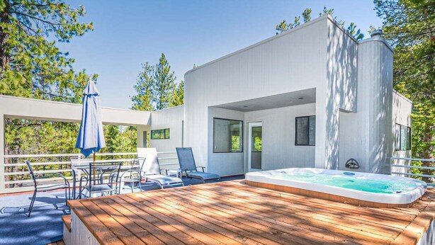 A modern outdoor deck with a hot tub, wooden furniture, and a white building with large windows.