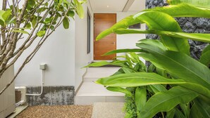 A modern building entrance with a wooden door, a stone wall, and lush green plants. - Exclusive Magnum Villa With Private Pool (Si Sunthon)