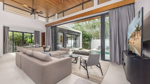 A modern living room with a large sofa, a coffee table, and a flat-screen TV. There is a dining area with a table and chairs, and a view of a swimming pool and greenery outside. - Exclusive Magnum Villa With Private Pool (Si Sunthon)