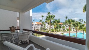 A balcony with wicker furniture, a glass table, and a view of a resort with palm trees and a pool.
