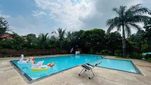 A swimming pool with two swan inflatable floats, a lounge chair, and palm trees in the background. - RKS RESORT KARJAT (Karjat)