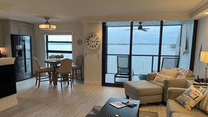 Amplia sala de estar con una mesa de comedor y sillas, un sofá y una mesa de centro. Un ventanal ofrece vista al agua.