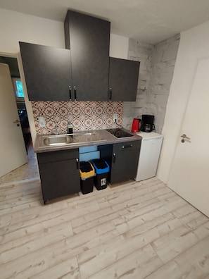 A compact kitchen with dark cabinets, a patterned backsplash, a sink, a coffee maker, and two trash bins.