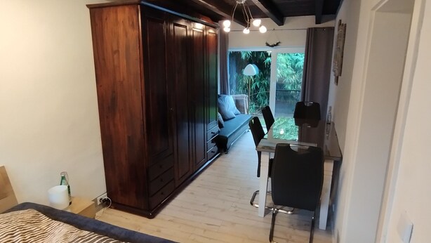 A bedroom with a bed, a wooden wardrobe, a desk with a chair, and a view of greenery outside.