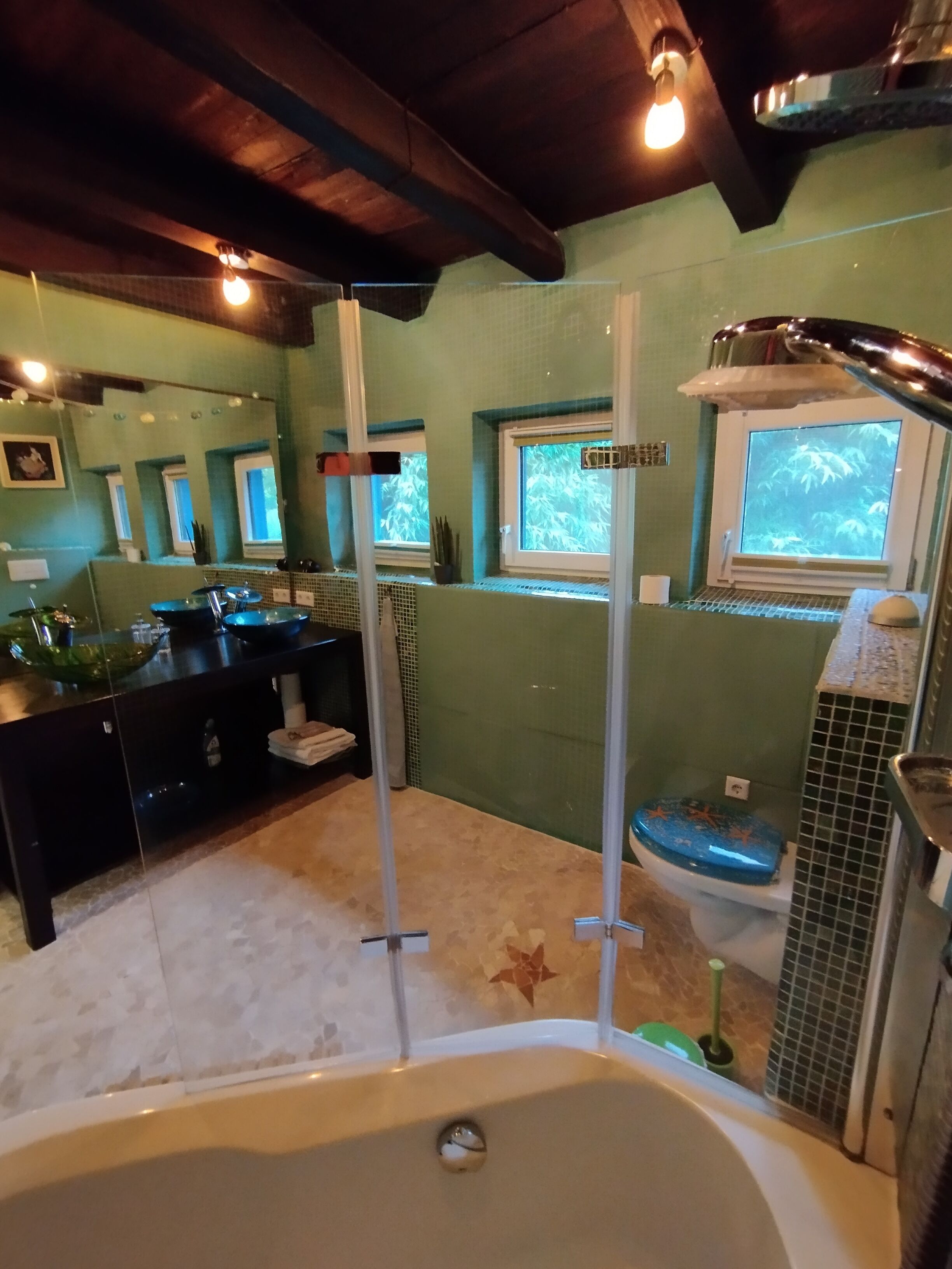 Une salle de bain avec une baignoire, des toilettes et un lavabo. Elle est dotée de fenêtres donnant sur des arbres et d’un espace douche séparé par une porte en verre.