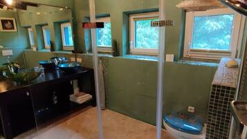 Une salle de bain avec une baignoire, des toilettes et un lavabo. Elle est dotée de fenêtres donnant sur des arbres et d’un espace douche séparé par une porte en verre.