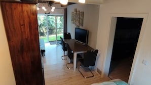 A bedroom with a bed, a wooden wardrobe, a desk with a chair, a television, and a view of a garden through a glass door.