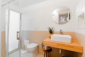 Une salle de bain avec des toilettes blanches, un meuble-lavabo en bois avec un lavabo blanc, un miroir rond et une plante en pot.