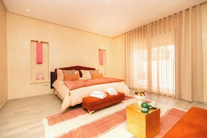 A modern bedroom with a large bed, a wooden bench, a small table with a vase, and sheer curtains. - Villa Stevana (Oulad Hassoune)