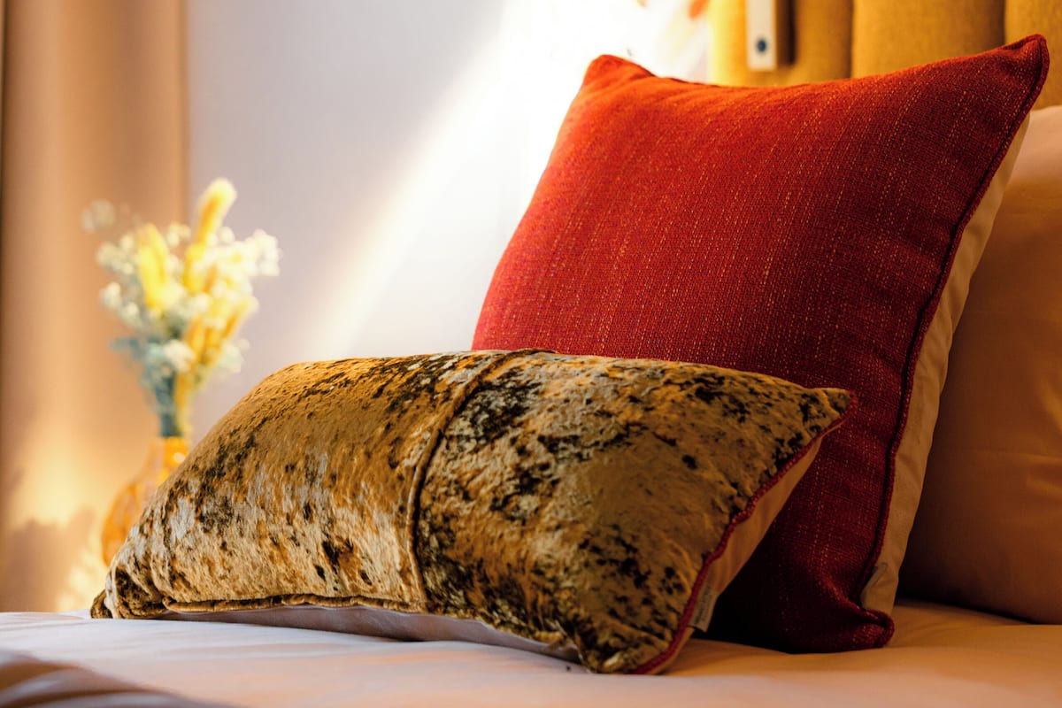 Un lit avec un oreiller rouge et un oreiller texturé, un oreiller jaune, et un mur à motifs en arrière-plan.