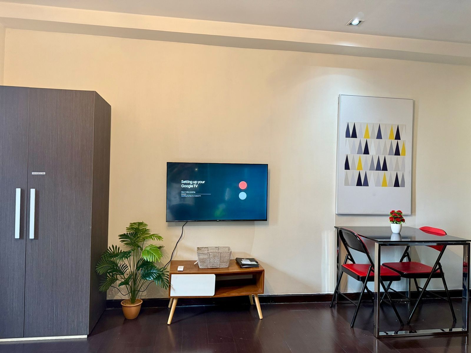A modern room with a flat-screen TV mounted on the wall, a wooden side table with a plant, a desk with a chair, and a framed abstract art piece.