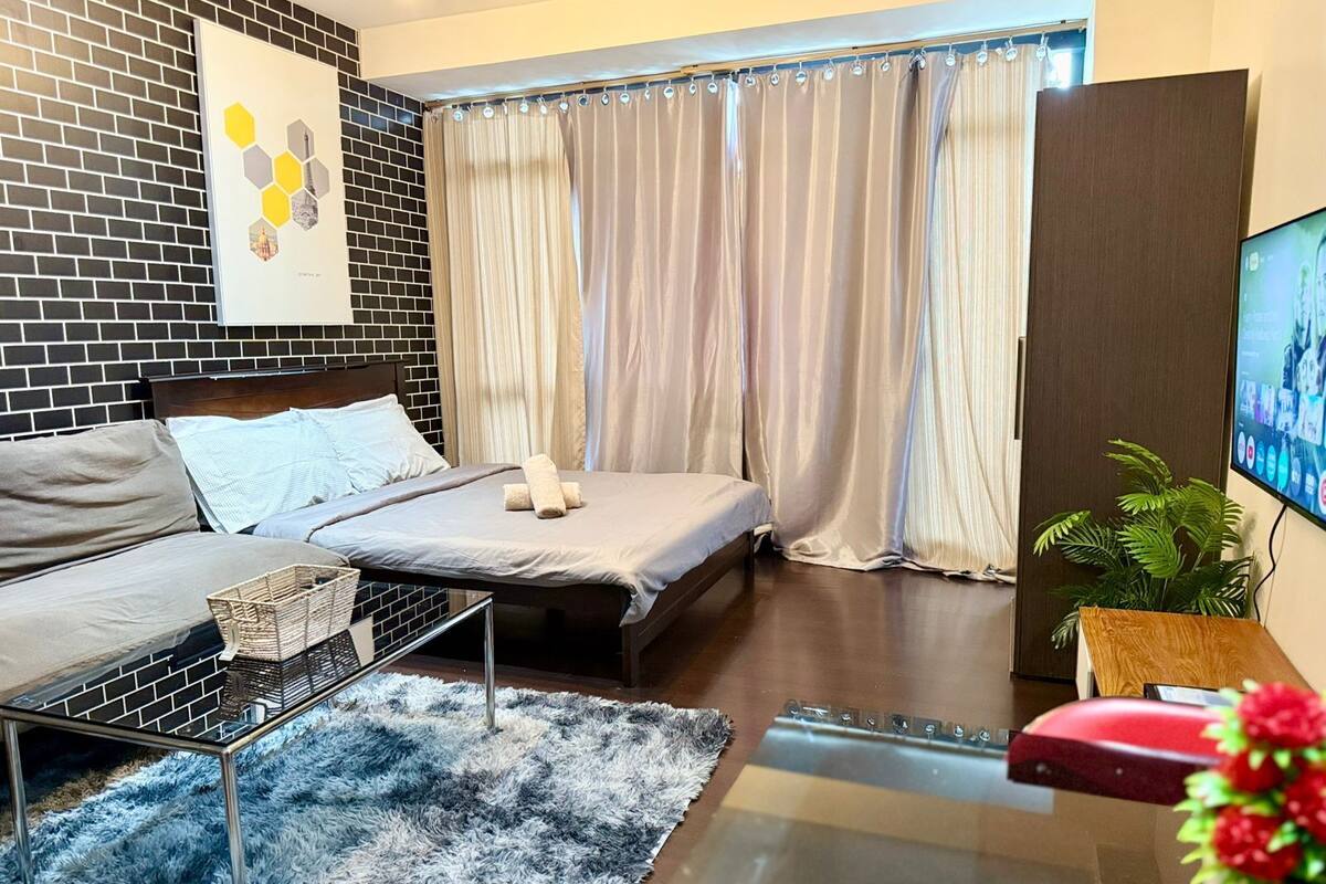 A modern hotel room with a brick-patterned wall, a bed with a grey headboard, a grey sofa, a small table with a basket, a fluffy blue rug, a wooden side table with a plant, and a TV mounted on the wall.