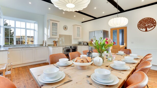Une cuisine moderne avec des meubles blancs, une table à manger dressée pour six personnes et une grande fenêtre laissant entrer la lumière naturelle.