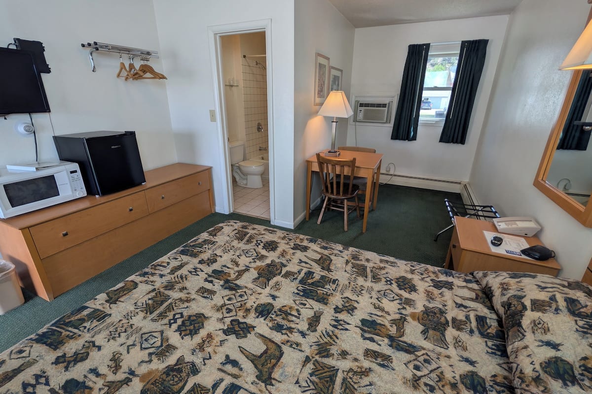 Une chambre d’hôtel équipée d’un lit, d’un bureau, d’une chaise, d’un four à micro-ondes, d’un radiateur, d’une lampe, d’un miroir et d’une fenêtre avec des rideaux.