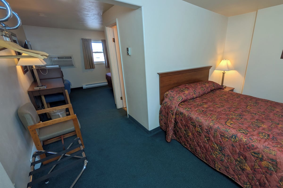 Une chambre d’hôtel de taille réduite, comprenant un lit simple, un bureau avec une chaise, une petite table, une échelle et une fenêtre avec des rideaux.