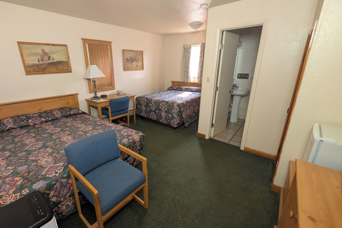 Une chambre d’hôtel avec deux lits, un bureau, une chaise, une salle de bain avec un lavabo et des toilettes, et deux tableaux accrochés au mur.