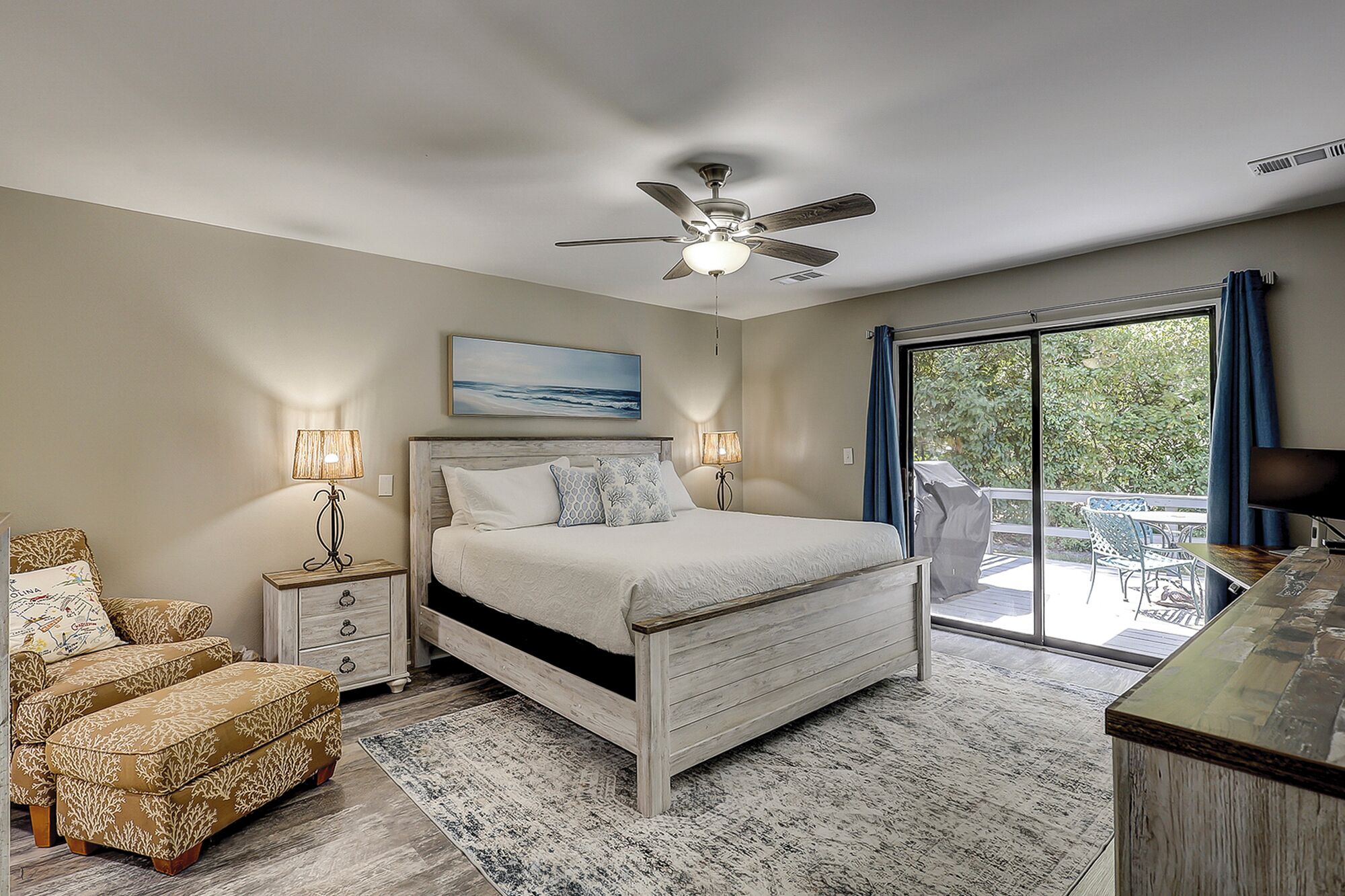 A bedroom with a bed, bedside lamps, a ceiling fan, a view of the outdoors, and a dining area with a table and chairs.