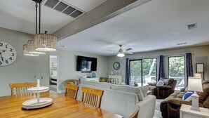A spacious living area with a wooden dining table, comfortable seating, a flat-screen TV, and large windows offering a view of the outdoors.