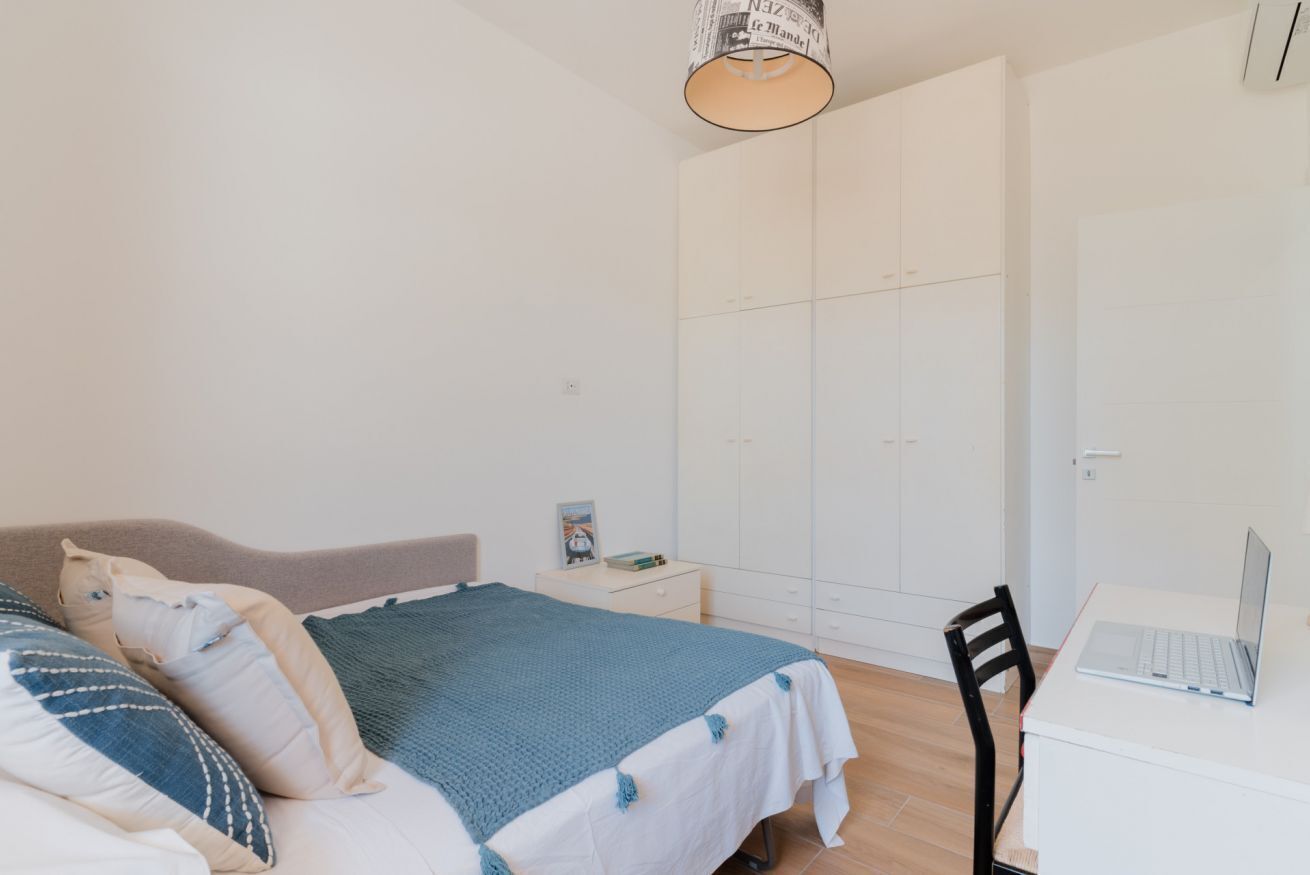 A bedroom with a bed, a desk with a laptop, a wardrobe, and a bedside table with a lamp.