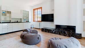 Een moderne keuken met witte kasten, een zwart aanrecht en een groot raam. In de woonkamer staan twee grijze pluin fauteuils.