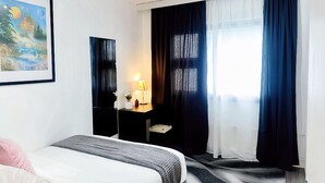 Et moderne hotelværelse med seng, skrivebord med lampe, et maleri på væggen og store vinduer med gardiner.