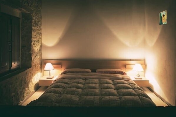 Un dormitorio con pared de piedra, una cama con colcha, dos lámparas de noche y un cuadro en la pared.