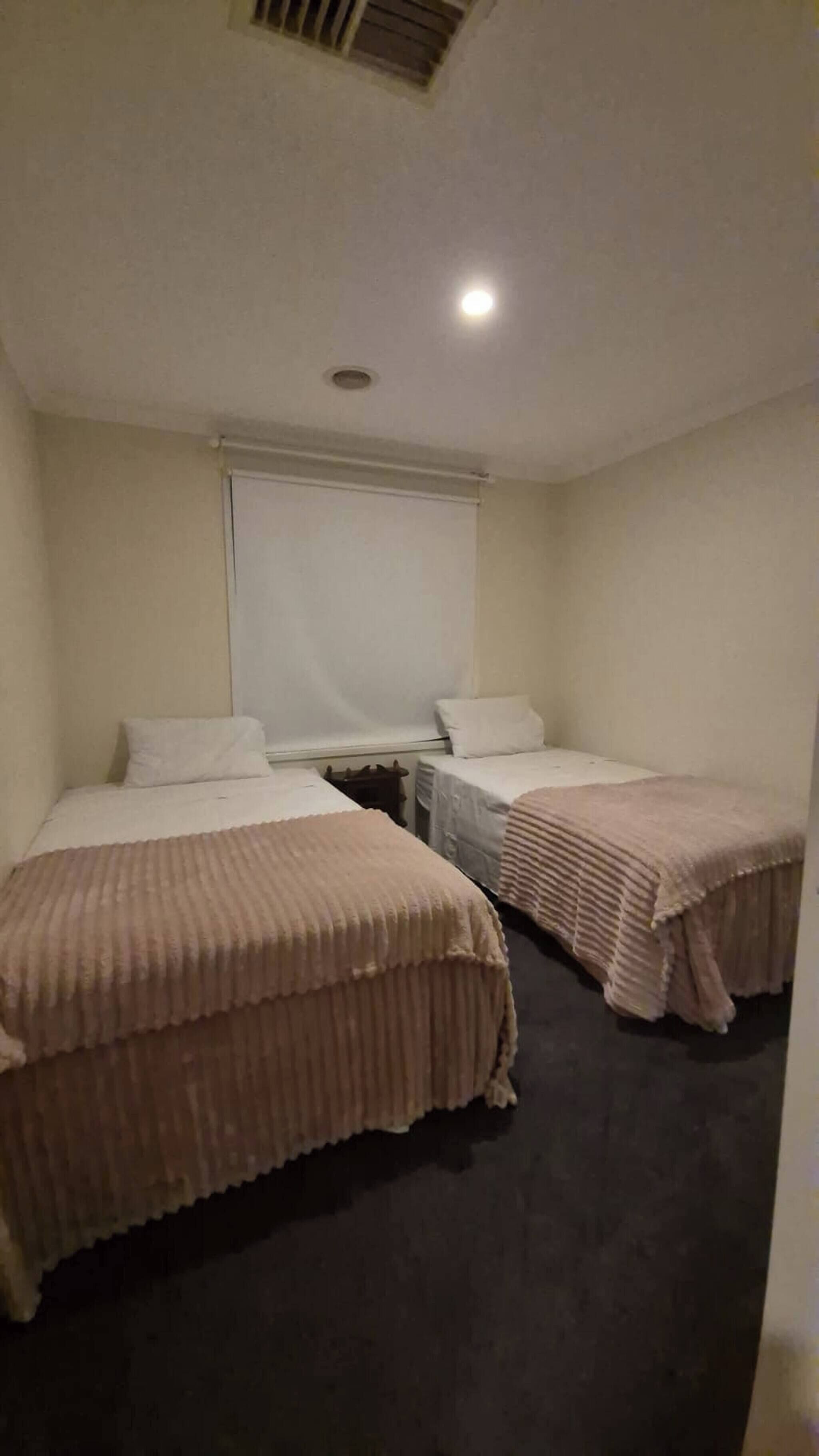 Una habitación pequeña y tenuemente iluminada, con dos camas individuales, una mesita y una ventana con persiana.
